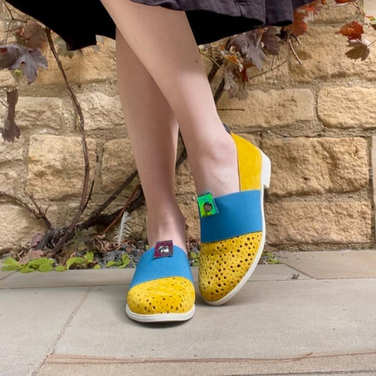 Yellow and blue shoes worn by a person standing on a stone pavement with a stone wall background.